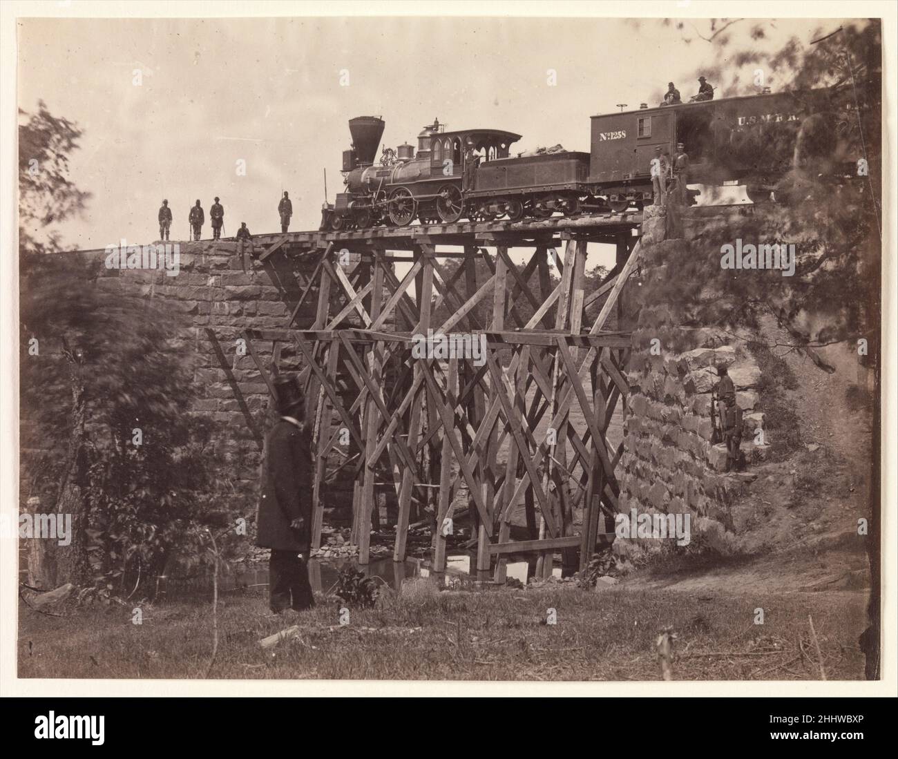 Brücke auf der Orange und Alexandria Rail Road, Wie von den Armeeingenieuren unter Oberst Herman Haupt repariert 1865 Andrew Joseph Russell American Diese Ansicht von Russell dokumentiert den Motor Fire Fly, der die Stabilität einer neuen hölzernen Böstelbrücke testet, die von den Ingenieuren der United States Military Railroad schnell gebaut wurde, um eine von den Konföderierten zerstörte Mauerwerksbrücke zu ersetzen. Auf dem Kastenwagen hinter dem Motor sind zwei bewaffnete Wachen; andere befinden sich auf den Gleisen und dem Brückenfundament. Die oben gehattete Figur im Vordergrund ist nicht identifiziert, aber er kann der Fotograf selbst oder eine zivile Eisenbahn sein Stockfoto