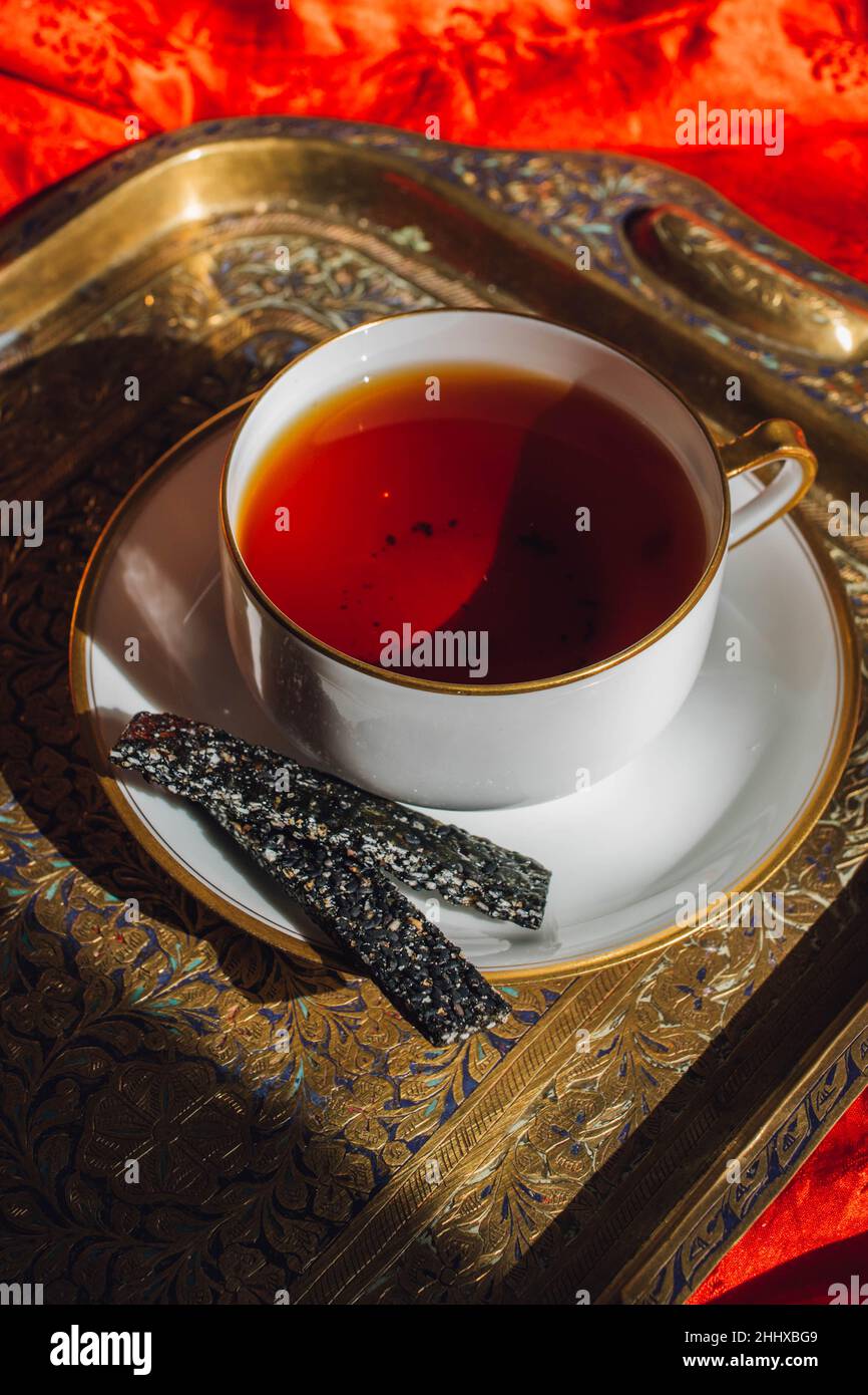 Weiße Teetasse mit heißem schwarzem Tee und schwarzen Sesamcrackern auf Messing versuchen gegen rote Seide Stockfoto