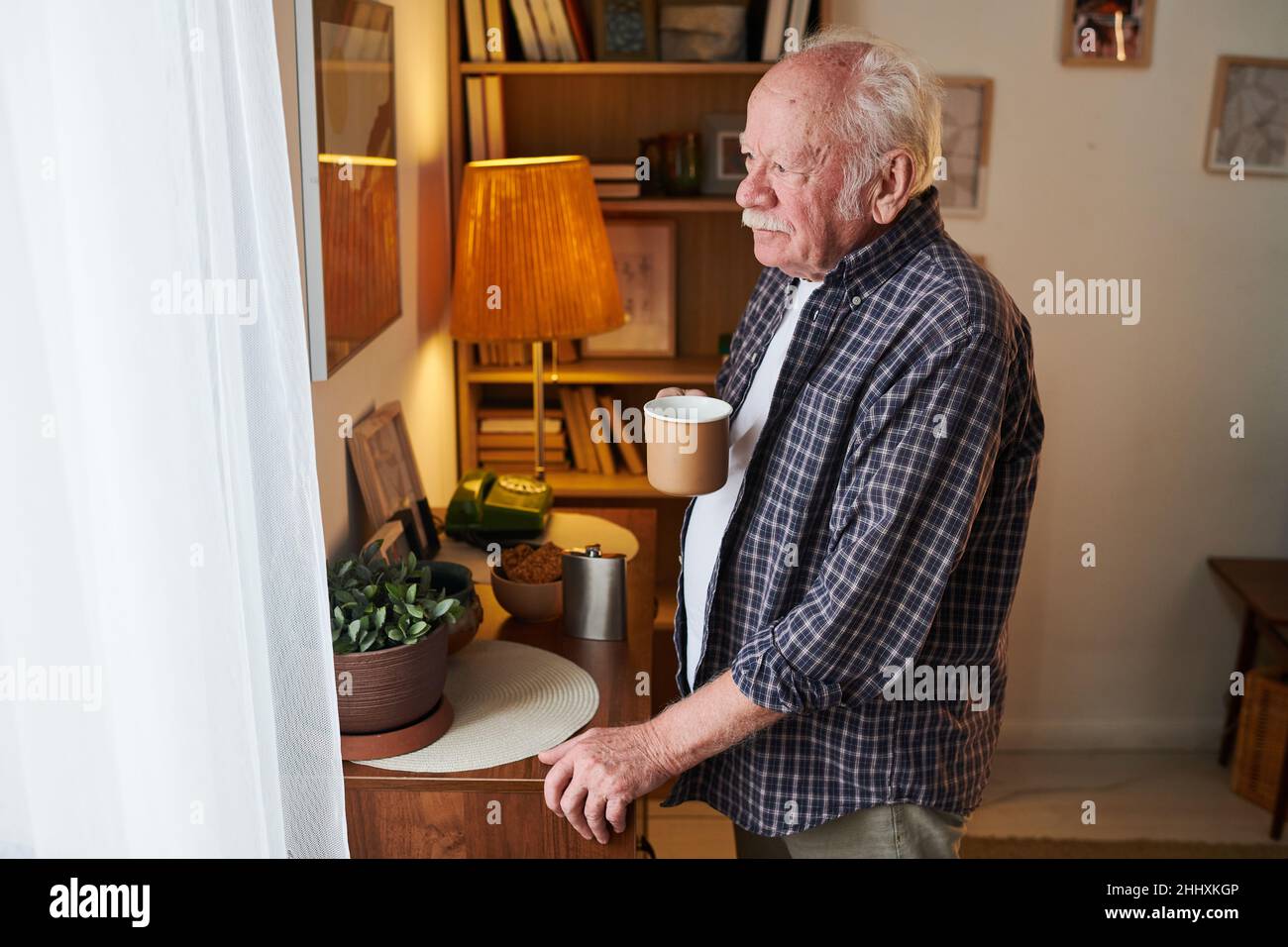Älterer Mann mit einer Tasse Tee, der vor dem Fenster mit weißen Nylonvorhängen stand und morgens nach draußen schaute Stockfoto
