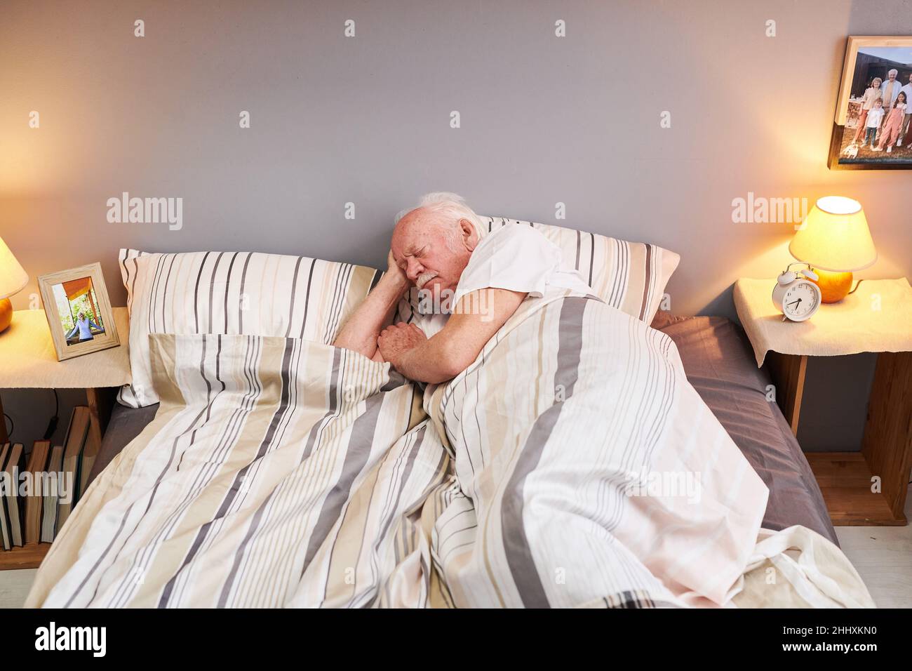 Ein älterer Mann, der den Kopf auf dem Kopfkissen hielt, während er unter einer gestreiften Decke auf dem Doppelbett im Schlafzimmer lag und schlief Stockfoto