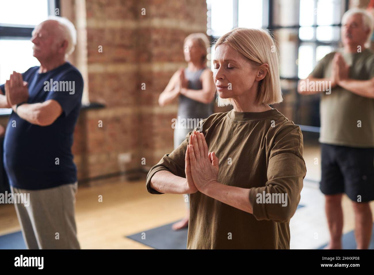 Eine Gruppe von reifen Männern und Frauen, die die Hände auf der Brust zusammenhalten, während sie Yoga-Übungen im Fitnessstudio praktizieren Stockfoto