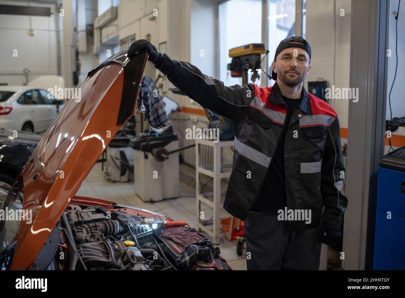 Junger männlicher Mechaniker in Arbeitskleidung, der die Abdeckung der Motorhaube öffnet, während er in der Garage oder in der Werkstatt neben dem Fahrzeug steht Stockfoto