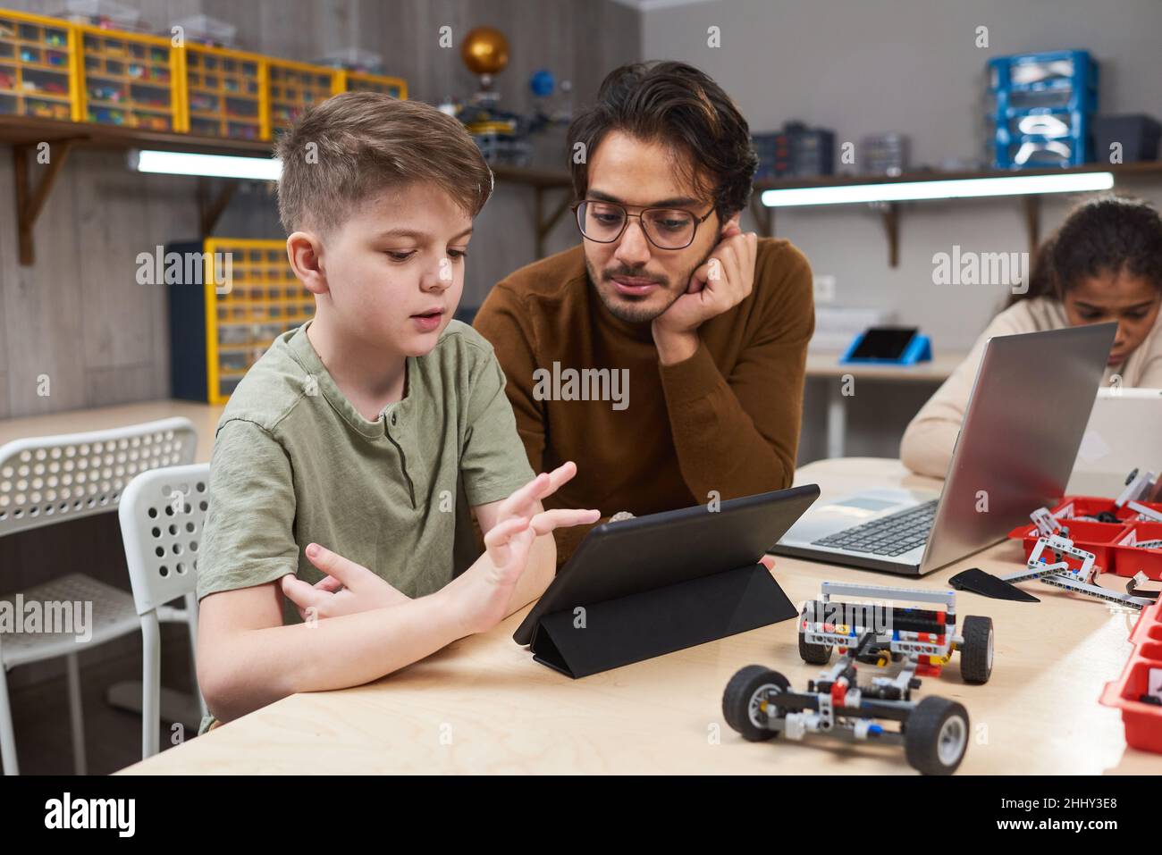 Junge mit digitalem Tablet während er mit dem Lehrer am Tisch saß, steuernde er den Roboter mit seinem Computer beim Roboterunterricht Stockfoto