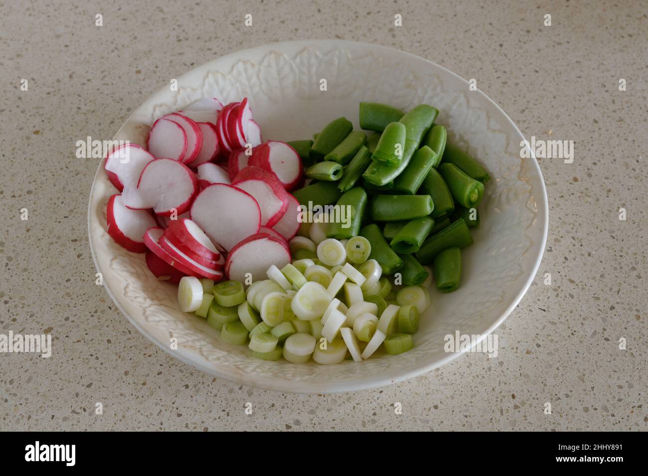 Frische Salatzutaten - Rettich, Frühlingszwiebeln, Zuckererbsen, die in einer weißen Schüssel auf beigem Hintergrund gehackt wurden Stockfoto
