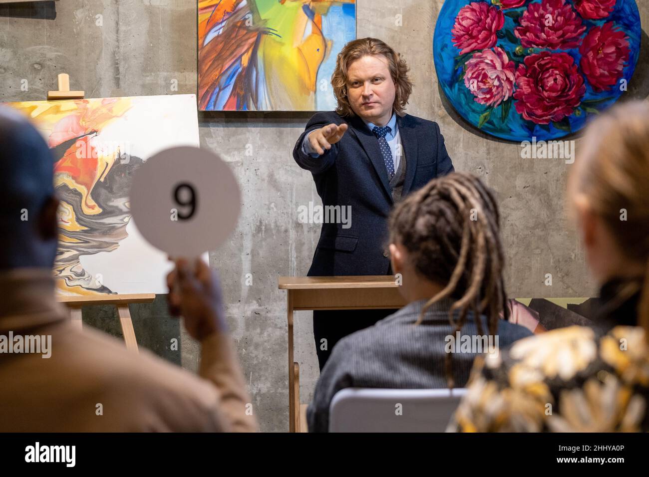Reifer Mann im Anzug zeigt auf Mann mit Tablet, Menschen kaufen Gemälde von berühmten Künstlern während der Auktion in der Galerie Stockfoto