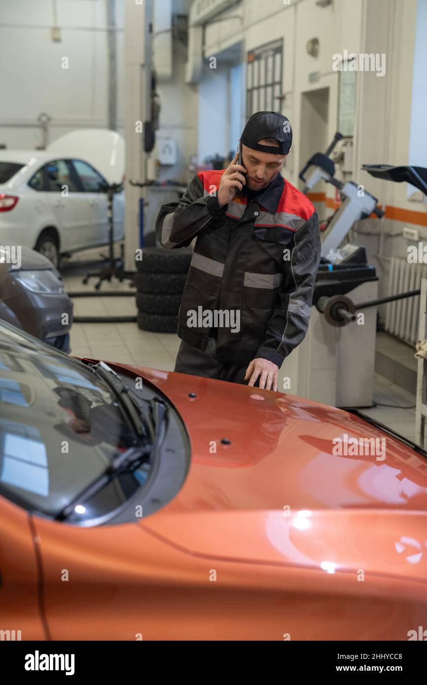 Ein junger, selbstbewusster Mechaniker, der mit dem Kunden auf dem Mobiltelefon spricht, während er nach der Überprüfung und Reparatur mit dem Auto steht Stockfoto