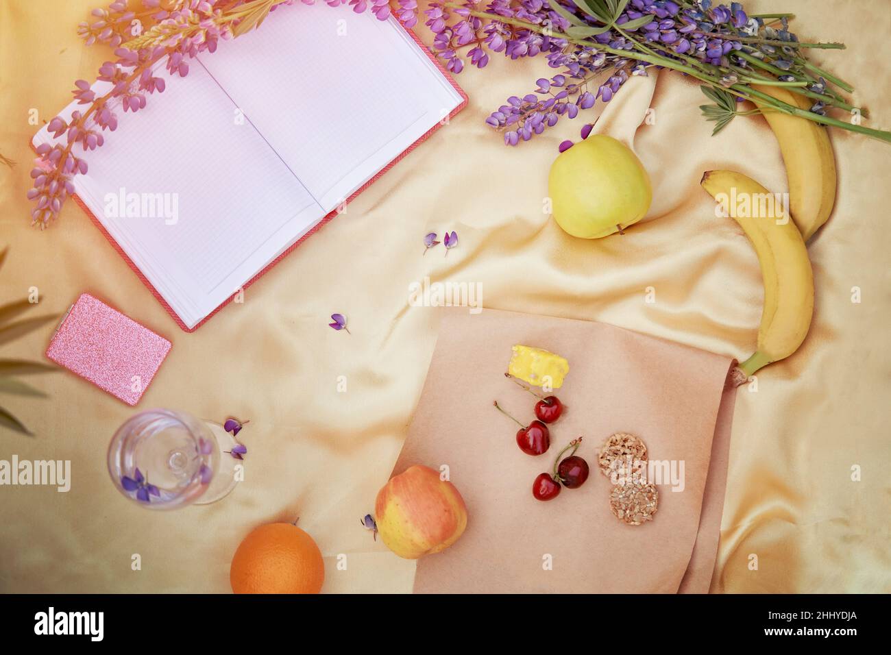 Offenes Notizbuch mit Weinglas, Lupinen, lila Blüten, Apfel, Bananen, Kirsche, Halva, Kekse auf goldenem Hintergrund. Picknick im Sommer. Für Text platzieren. Nähe zur Natur, Selbsterkennungskonzept Stockfoto