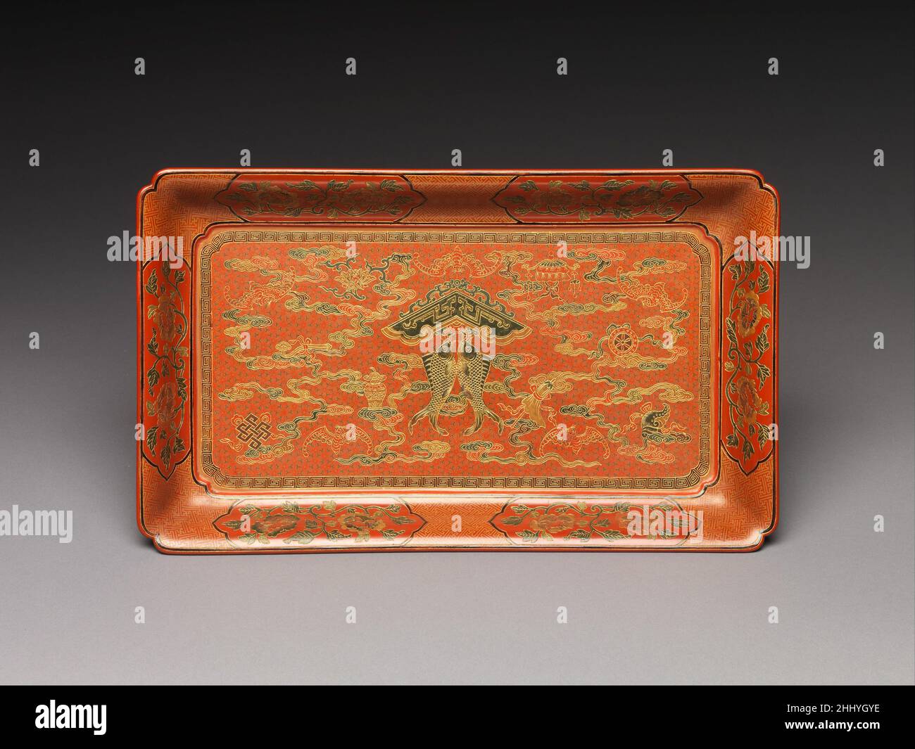 Rechteckiges Tablett mit Fischspiel, Schätzen und Wolken China die beiden Fische auf diesem Tablett hängen an einem Anhänger in Form eines alten Steingimens, das Wünsche nach Glück und Fülle (yuqing) darstellt. Der Zwillingsfisch ist auch einer der acht buddhistischen Schätze – die anderen sieben sind im Vordergrund angeordnet: Ein Rad, eine Muschelschale, ein Regenschirm, ein Sonnenschirm, eine Blume, Glas und Doppelknoten. Gezeigt mit Fledermäusen und Wolken, diese Symbole vermitteln glückverheißende Wünsche über religiöse Bedeutung hinaus.. Rechteckiges Tablett mit Fischspiel, Schätzen und Wolken 667293 Stockfoto