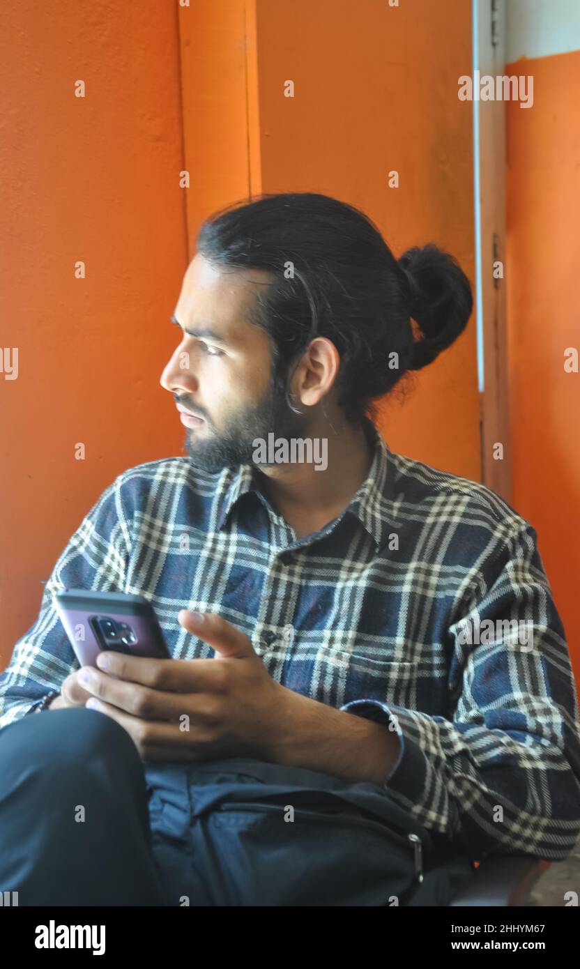 Selektiver Fokus eines gut aussehenden bärtigen Mannes mit Haarsträhne, der während der Benutzung seines Telefons seitwärts schaut, während er drinnen sitzt Stockfoto