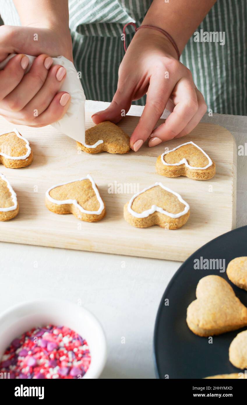 Damenhände schmücken Ready Cookies mit Glasur. Der Prozess der Herstellung von Urlaubsklätzchen zum Valentinstag. Mit Liebe zu Hause backen. Vertikale Orientatio Stockfoto