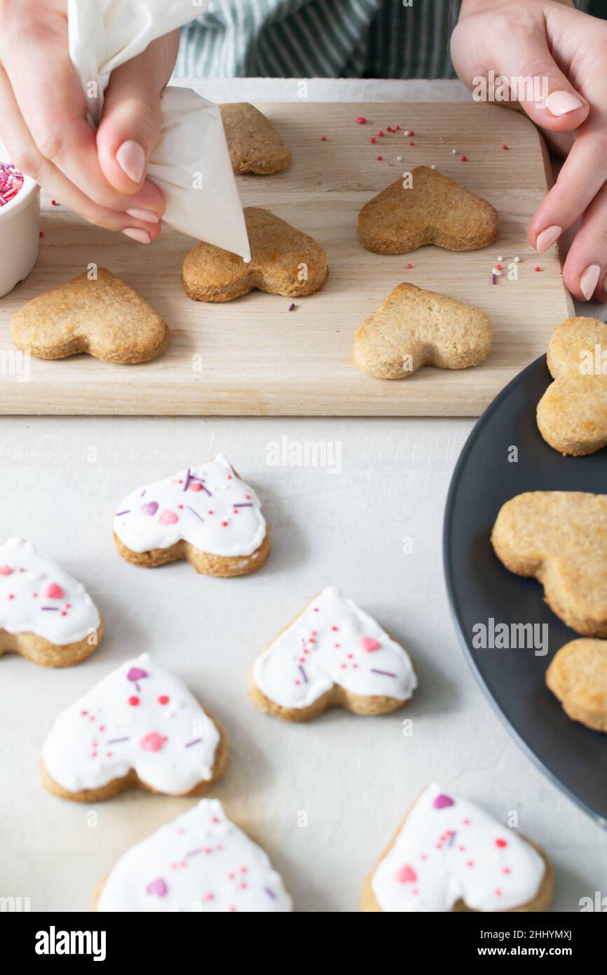 Damenhände schmücken Ready Cookies mit Glasur. Der Prozess der Herstellung von Urlaubsklätzchen zum Valentinstag. Mit Liebe zu Hause backen. Vertikale Orientatio Stockfoto