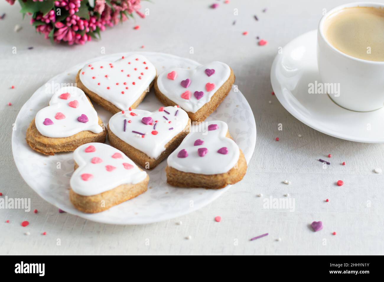 Lebkuchen mit weißer Glasur bedeckt und auf einer hellen Untertasse mit einer Tasse Espresso und Blumen im Hintergrund bestreuen. Das Konzept des Feierns Stockfoto