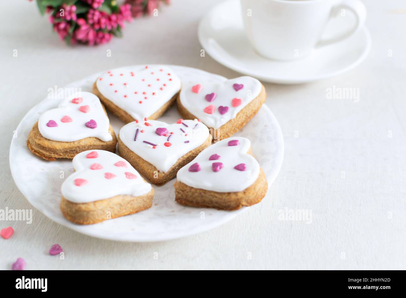 Lebkuchen mit weißer Glasur bedeckt und auf einer hellen Untertasse mit einer Tasse Espresso und Blumen im Hintergrund bestreuen. Das Konzept des Feierns Stockfoto