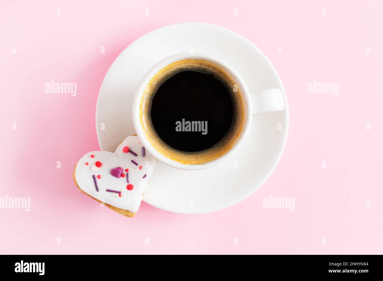 Weiße Tasse und Untertasse mit Espresso und Lebkuchen mit weißer Glasur bedeckt und bestreuen auf einem rosa Hintergrund. Das Konzept der Feier zum Valentinstag Stockfoto