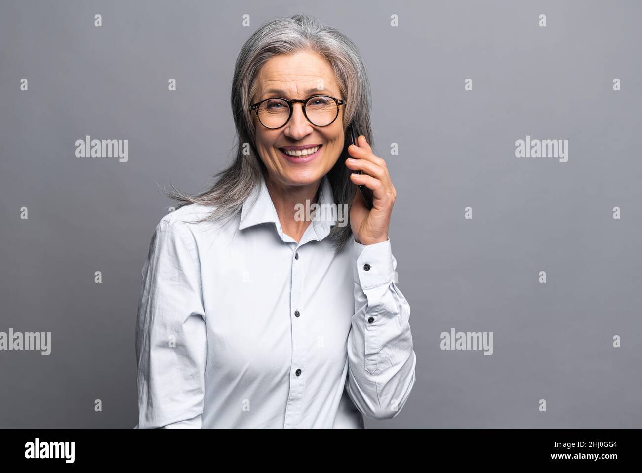 Lächelnde und positive Geschäftsfrau, die mit dem Smartphone über der grauen Wand spricht. Ältere Verkäuferin telefoniert. Reife weibliche Angestellte, die auf dem Mobiltelefon spricht Stockfoto