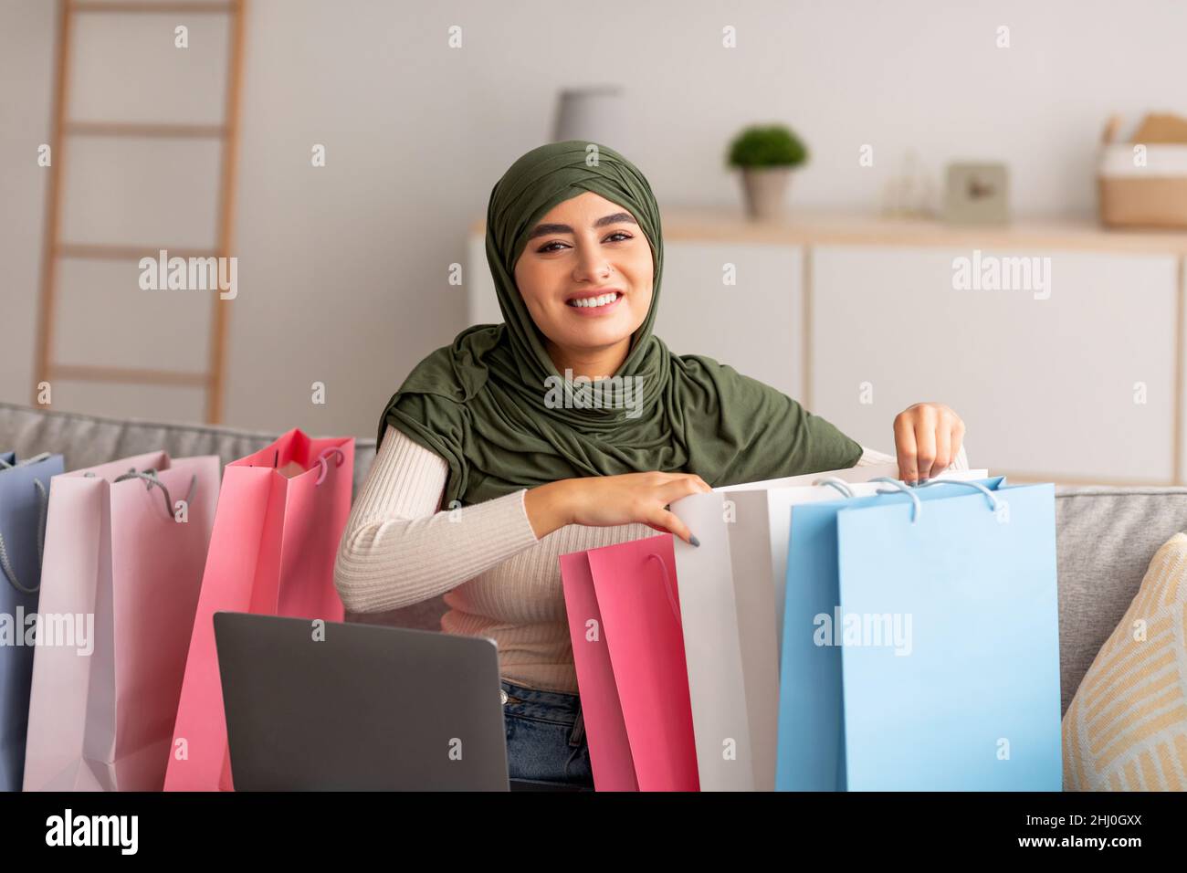 Glückliche arabische Frau in Hijab sitzt auf der Couch mit Laptop und hellen Einkaufstaschen, Waren online zu Hause kaufen Stockfoto