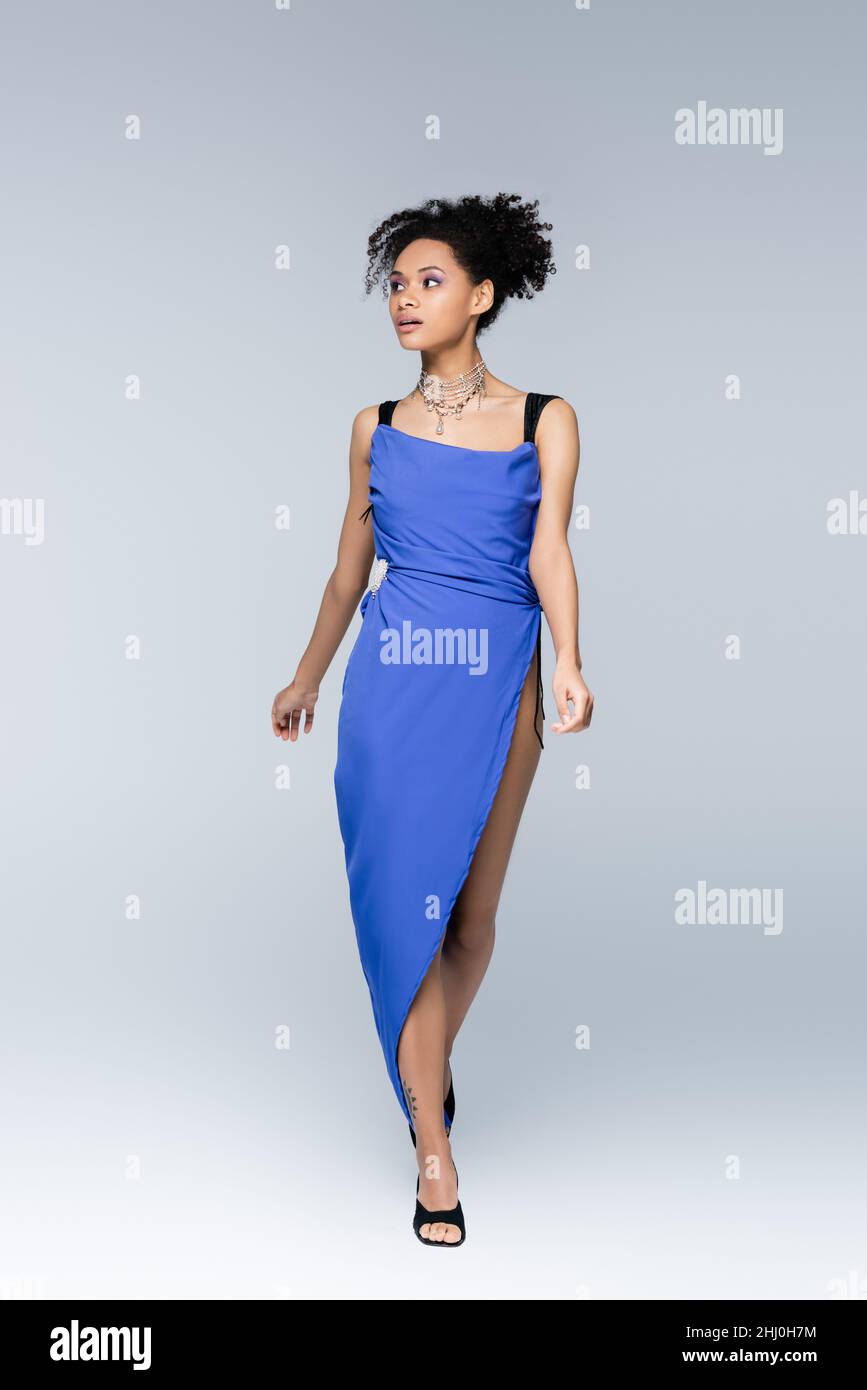 Volle Länge der lockigen afroamerikanischen Frau in hellblauem Kleid zu Fuß auf grau Stockfoto