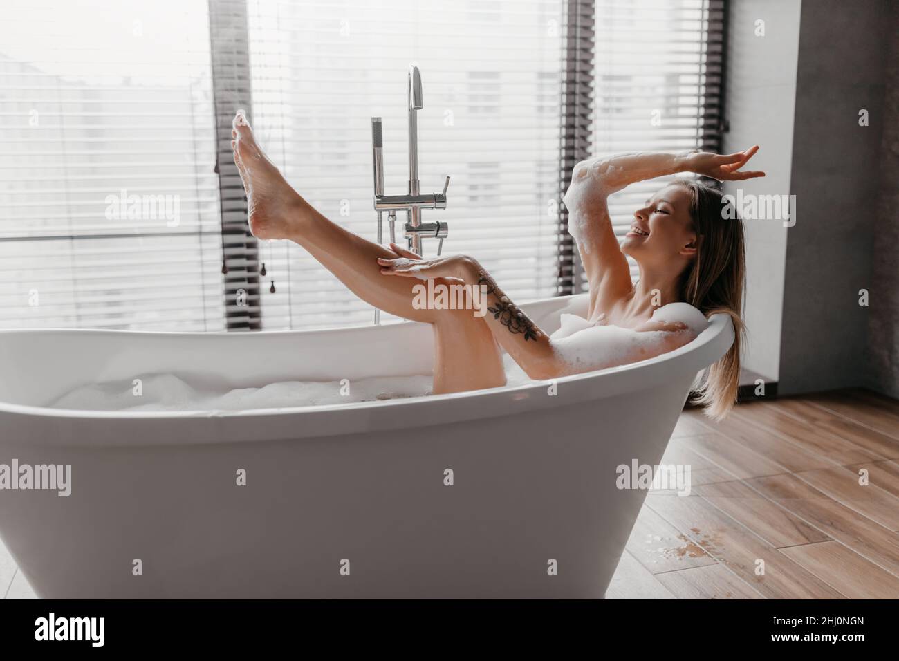 Home Spa und entspannen Sie am Wochenende. Hübsche Frau in der Badewanne mit Schaumstoff, das Bein hochhebend und genießt das Baden in der Badezimmereinrichtung Stockfoto