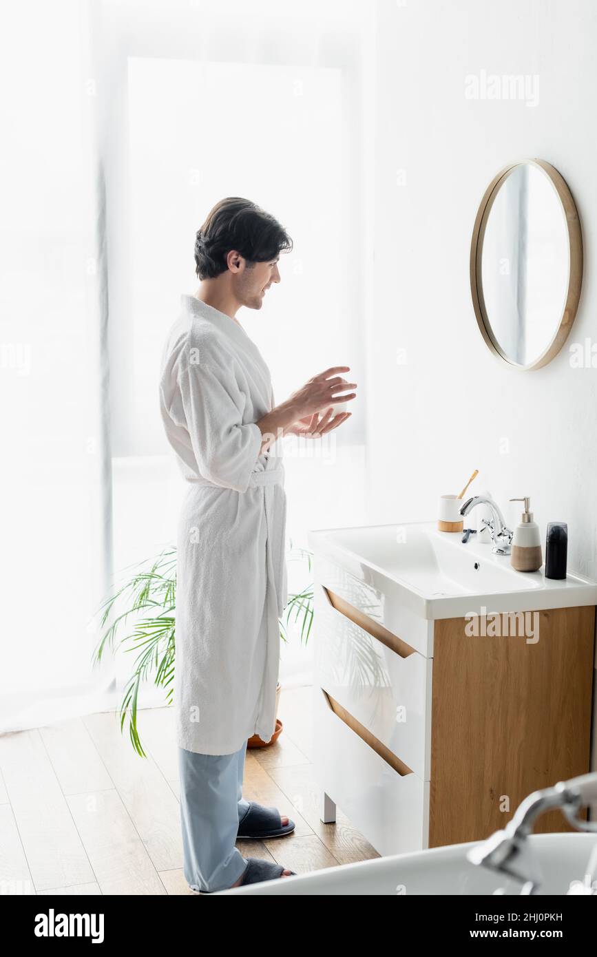 Blick auf den Mann in weißem Bademantel, der Kosmetikcreme neben dem Spiegel und Toilettenartikeln auf dem Waschbecken hält Stockfoto