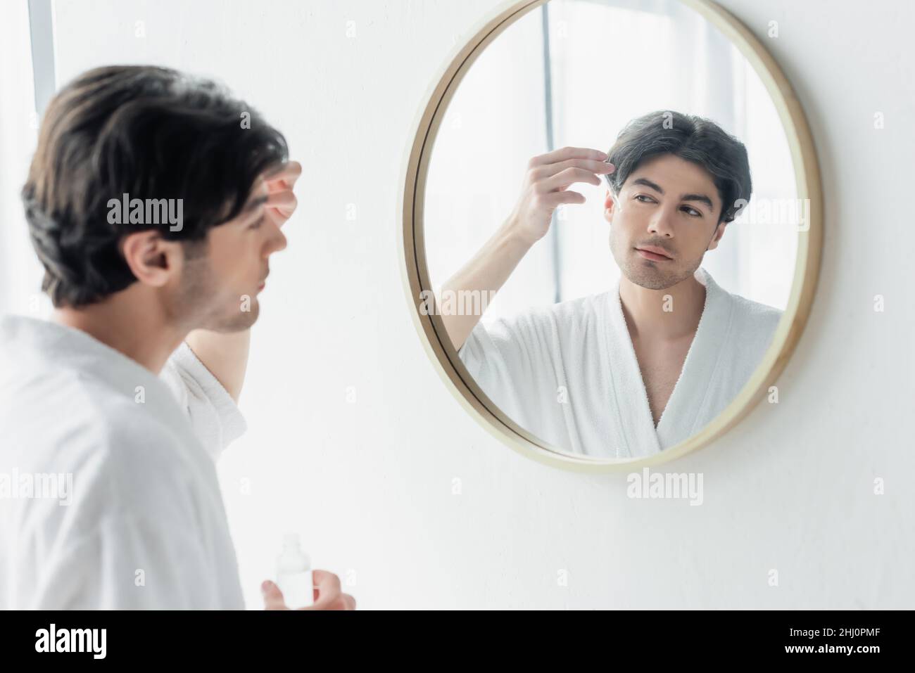 Junger Mann im weißen Bademantel, der kosmetisches Serum in der Nähe des Spiegels im Badezimmer anwendet Stockfoto