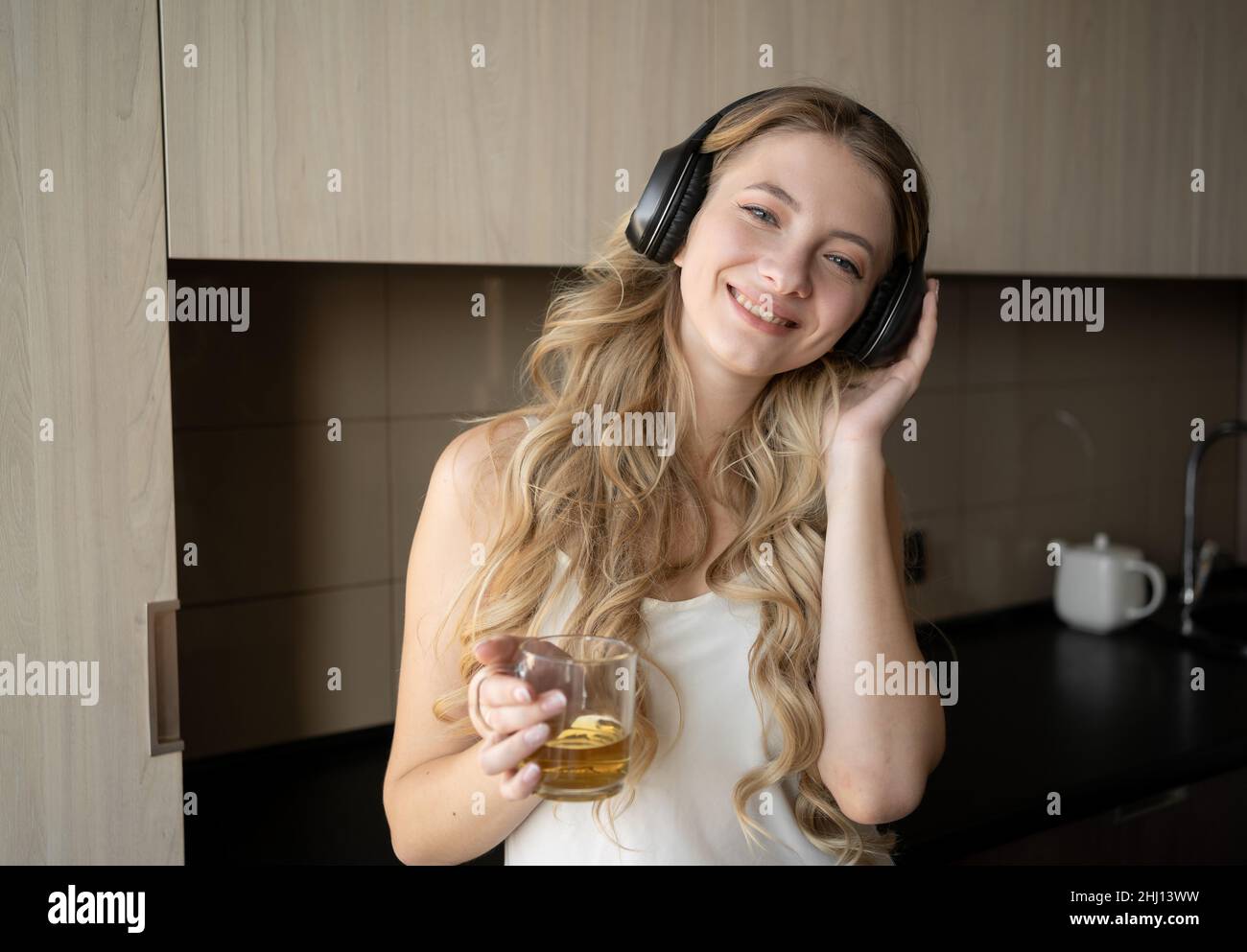 Die kaukasische Frau in einem weißen Hemd mit Kopfhörern auf dem Kopf tanzt morgens in der Küche zu Hause. Eine Frau hört Musik, während sie sie trinkt Stockfoto