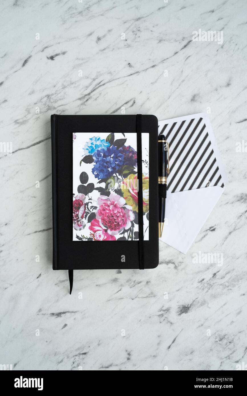 Schwarzer Notizblock in der Mitte des Marmortisches, neben einer hellen Karte mit Blumen, einem weißen Umschlag und einem stilvollen Stift Stockfoto