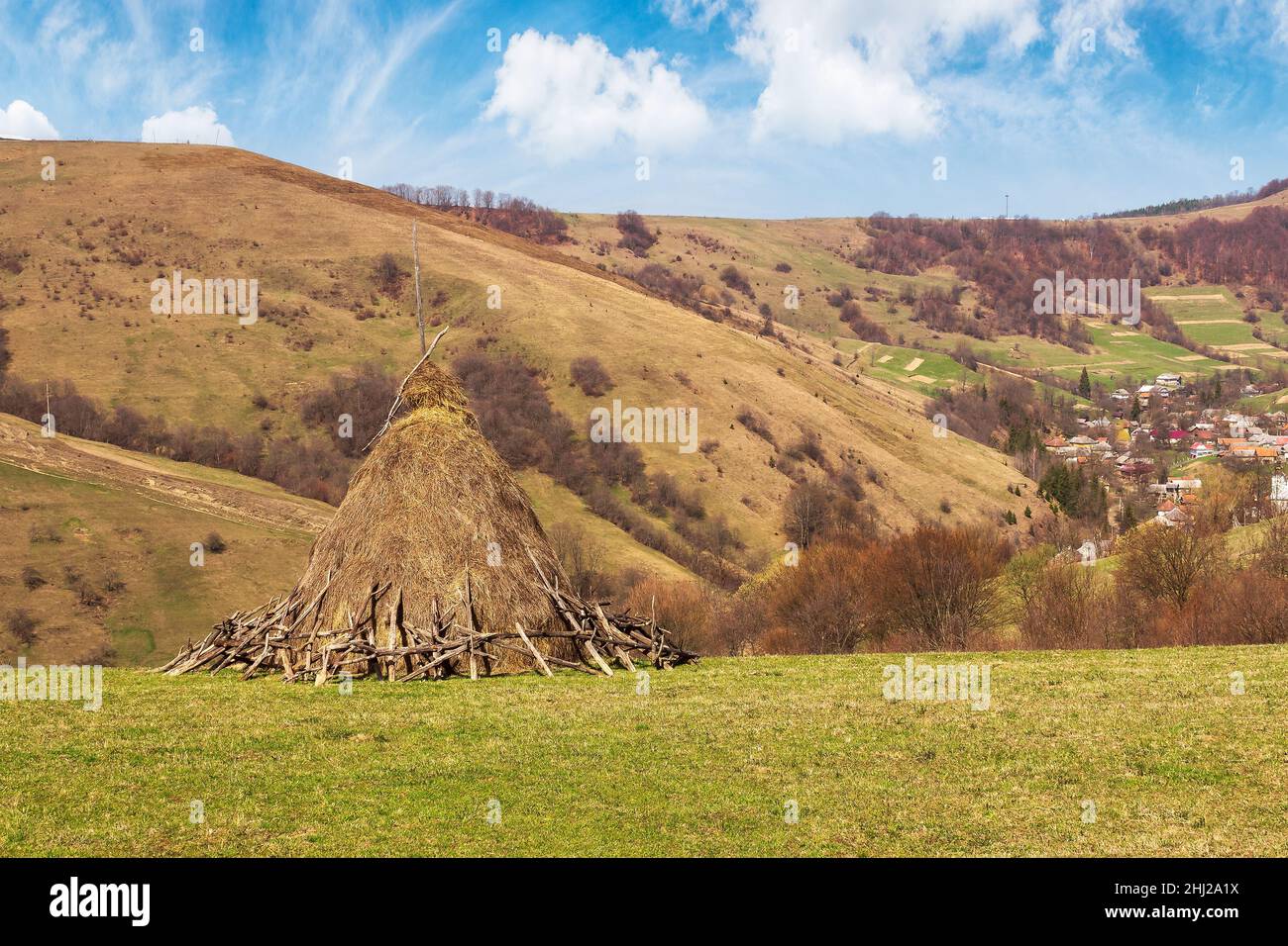 karpaten ländliche Landschaft im frühen Frühjahr. Heuhaufen auf dem grasbewachsenen Hügel, Dorf im Tal, Gipfel in der Ferne Stockfoto