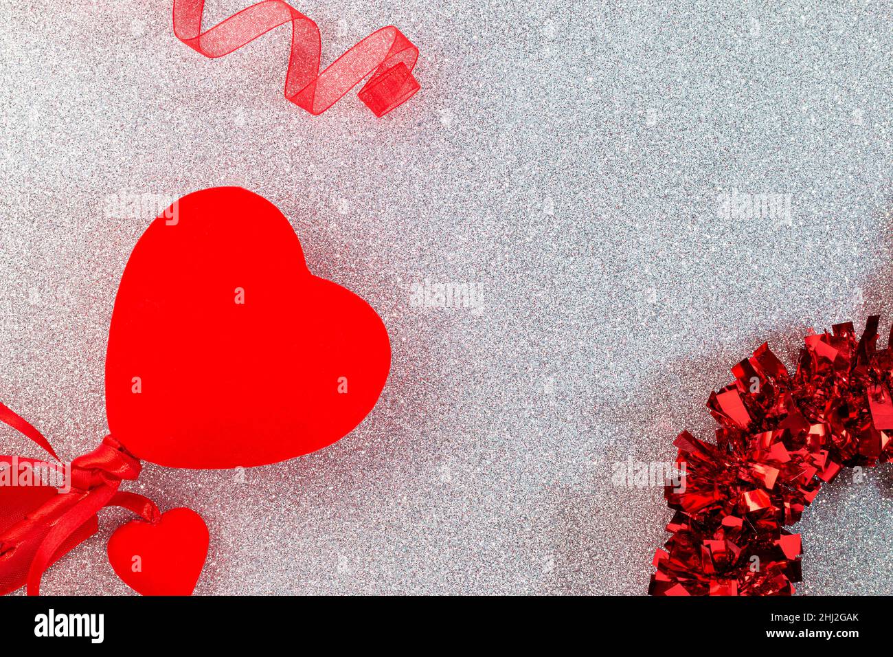 Auf einer silbernen Glitzeroberfläche sind rote Dekorationen wie ein Band, Herzen und eine Girlande zu sehen, im Rest des Bildes gibt es leeren Raum. Stockfoto