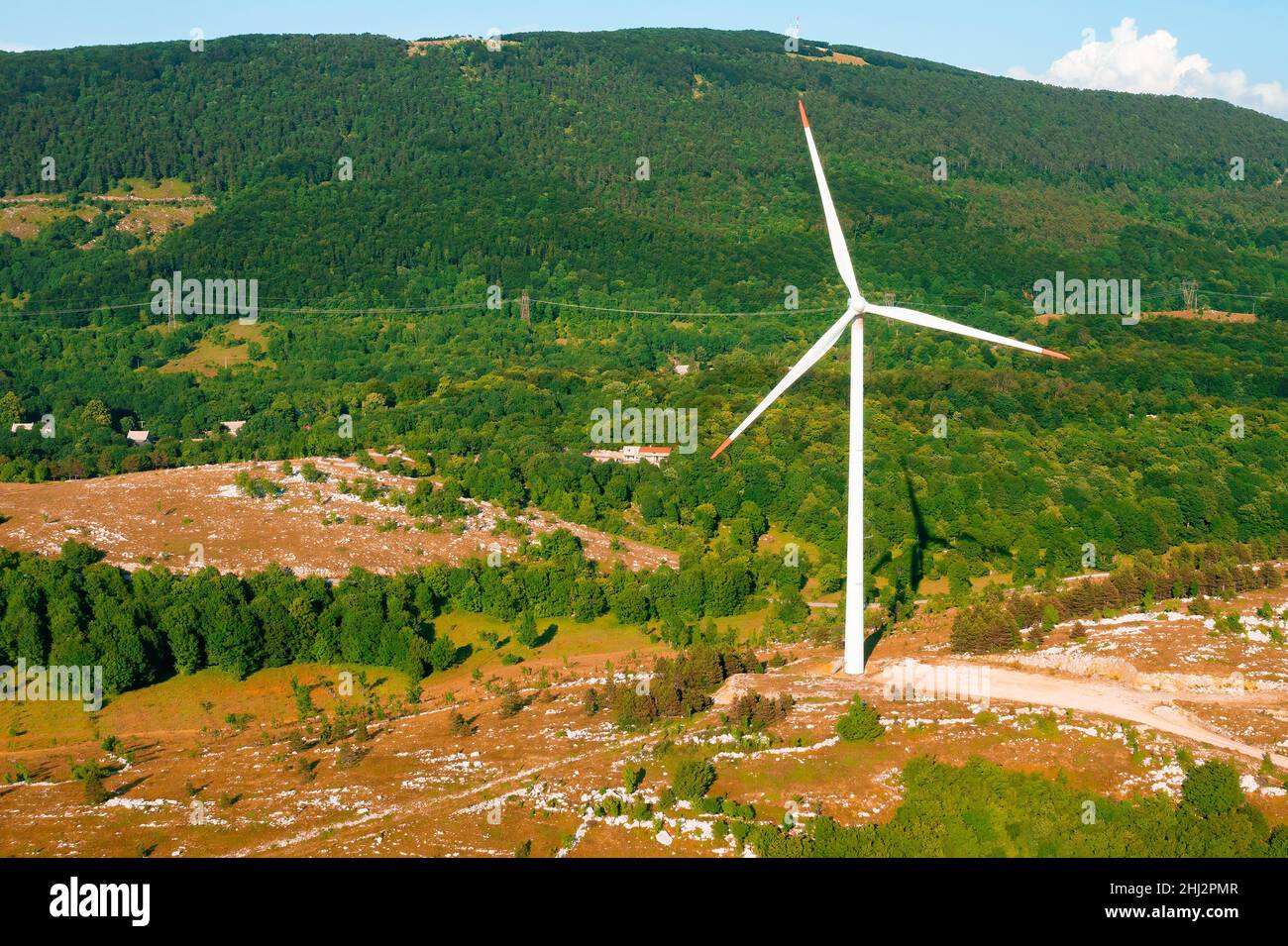 Der moderne windbetriebene Generator produziert grüne Energie gegen den großen Waldberg im Hochland bei sonniger Tagesansicht von der Drohne aus Stockfoto