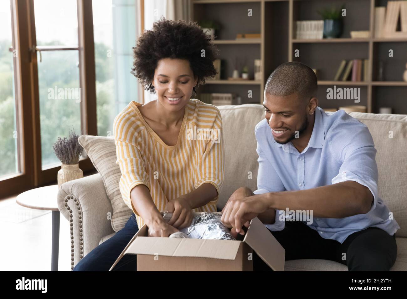 Glückliches afrikanisches Paar, das Paket auspackt, sitzt zu Hause auf dem Sofa Stockfoto