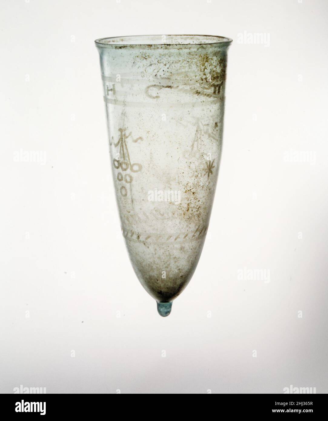 Glasbecher mit geschliffenen Verzierungen 4th–Anfang 5th Jahrhundert n. Chr. römisch lichtdurchlässig hellblau. Senkrechter Rand, abgesprungen und flach geschliffen, mit Ausbuchtung unten; konischer Körper mit seitlicher Verjüngerung sanft nach unten; Massiver hervorstehender Knopf auf der Unterseite.Radabrieb-Dekoration, bestehend aus zwei breiten horizontalen Linienbändern, die griechische Buchstaben mit einem diagonalen Band aus kurzen Linien umschließen, die den Anfang und/oder das Ende der Inschrift markieren; In der Mitte ein breiter Fries mit zwei stilisierten Trauben, die von einem T-Balken aufgehängt und von welligen Bändern flankiert wurden, die sich um die Seite mit zwei achtarmigen Sternen wechselten: belo Stockfoto