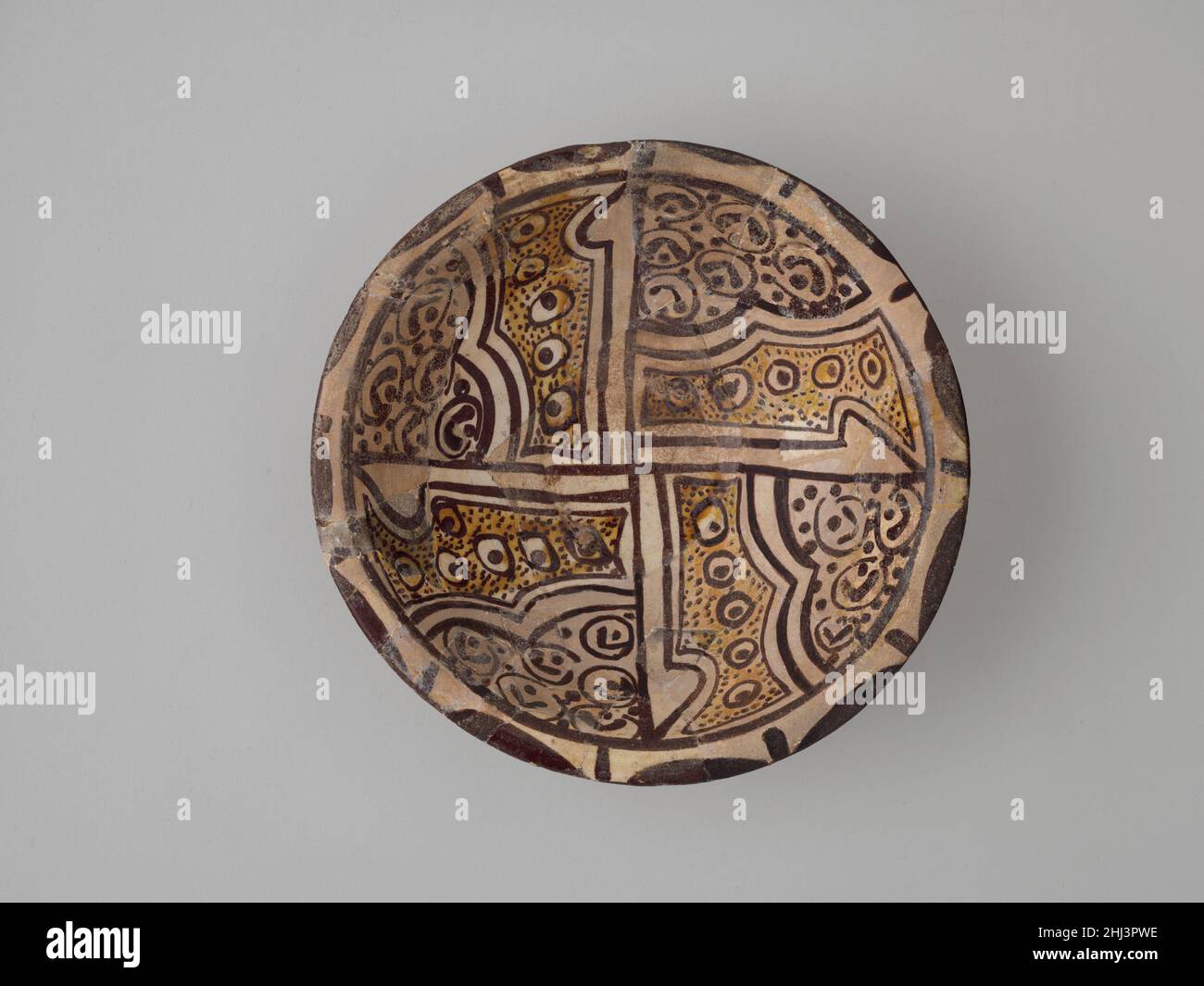 Imitation Bichrome Luster Bowl 9th Century die Dekoration dieser Schale imitiert eine Art von Luster Keramik, die in der Regel auf die Mitte des bis Ende des neunten Jahrhunderts datiert wird und sich durch die Verwendung von zwei Farbtönen einer bräunlich-goldenen Glasur auszeichnet. Statt jedoch mit der Glasur auf Metallbasis aus Glanzware hergestellt zu werden, ist diese Schale mit braunen und gelben Glasuren aus verdünntem Ton verziert. Sein Inneres ist durch vertikale Striche in Vierteln unterteilt; jeder Quadrant ist mit dem „Pfauenauge“-Muster verziert, das so häufig bei Glanzwaren dieser Periode zu finden ist. Imitation Bichrome Lustre Bowl. 9th Jahrhundert. Steingut; weiß Stockfoto