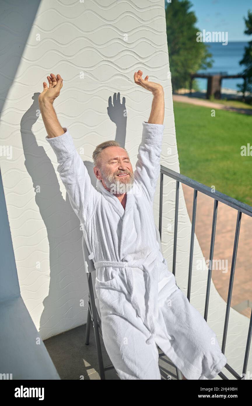 Grauhaariger Mann im weißen Bademantel, der im Sonnenschein sitzt Stockfoto