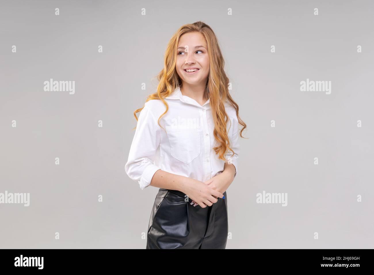 Junge blonde Frau, die in einem weißen Hemd in Relief gekleidet ist, steht entspannt und lächelt, dankbar für die Person, die ihr geholfen hat, isoliert auf grauem Hintergrund. Schöne kaukasische Mädchen niedlich suchen schüchtern Stockfoto