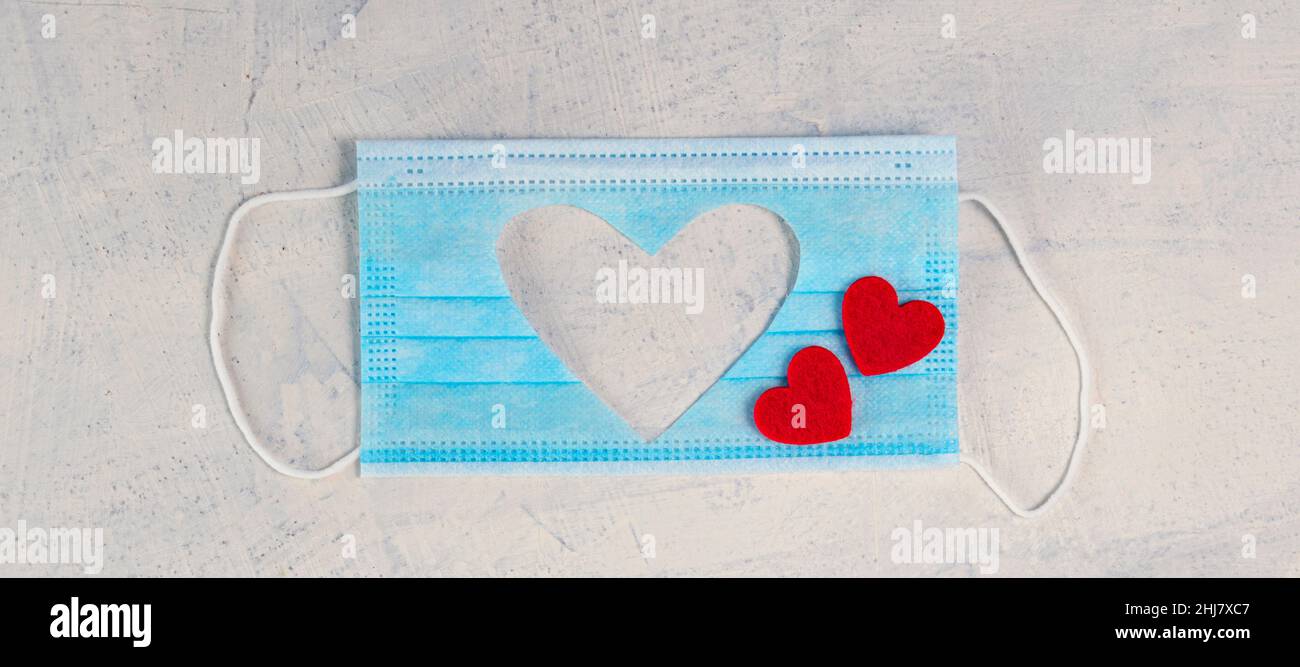 Banner mit medizinischer Maske mit ausgeschnittener Silhouette des Herzens und zwei roten Herzen auf weißem Stein Hintergrund. Valentinstag Hintergrund. Valentinstag-Konzept. Flach liegend. Draufsicht. Speicherplatz kopieren Stockfoto