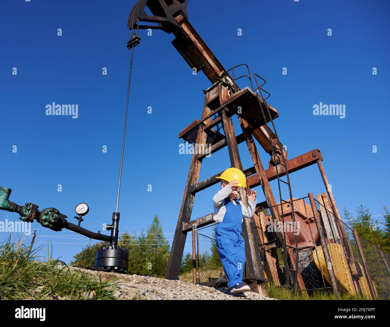 Low-Angle-Ansicht eines kleinen jungen Energietechnikers in der Nähe der Ölplattform. Boy in Overalls gegen den blauen Himmel, Petroleum-Pipeline und Pumpenheber für die Gewinnung von natürlichem Kraftstoff. Stockfoto
