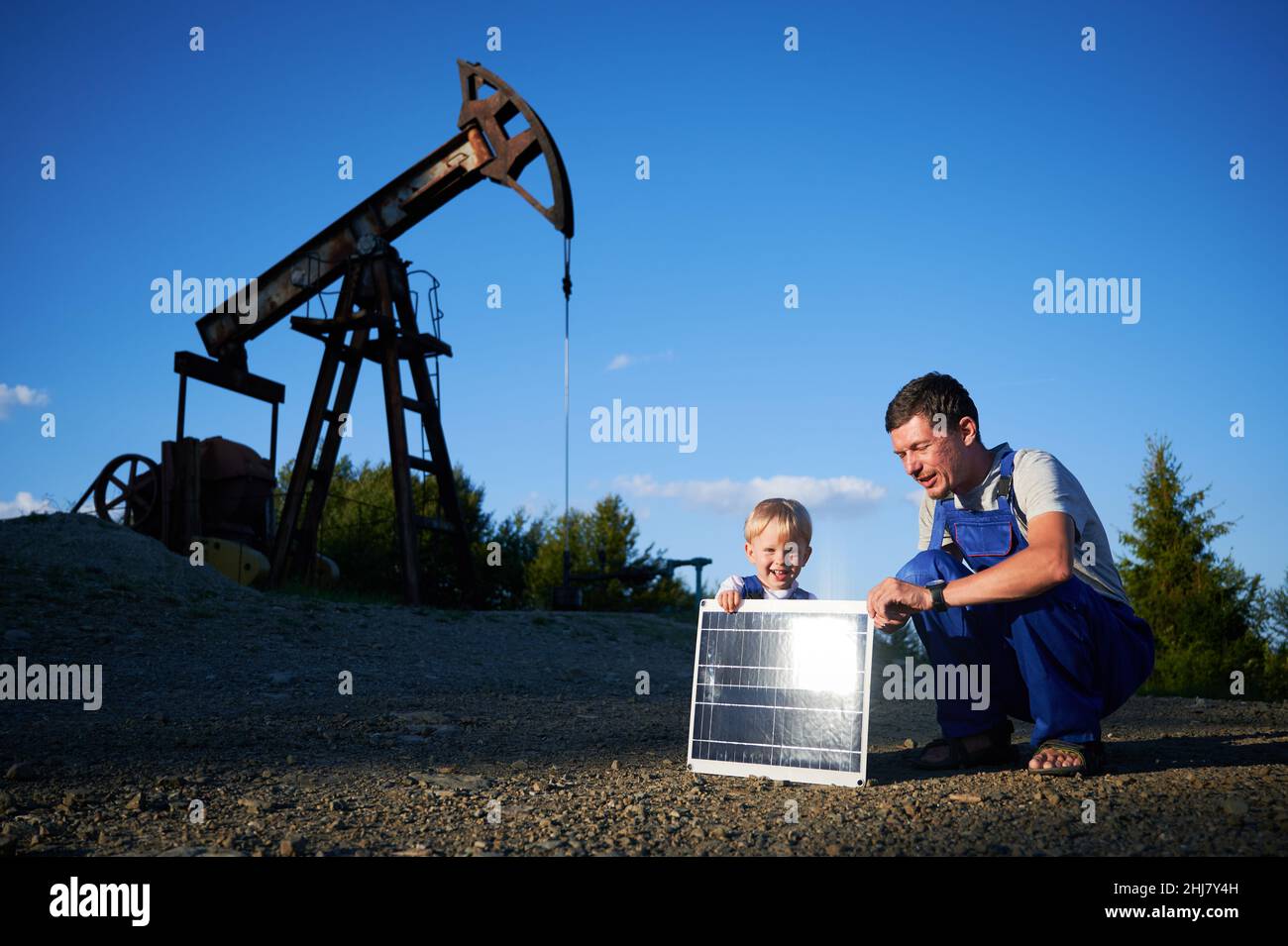 Erwachsener Mann und männliches Kind setzten sich auf Schotterplatz in der Nähe des Ölbrunnen. Sie halten Solarbatterie, Sonnenstrahlen von Solarpanel reflektiert. Zwei Varianten der Energieerzeugung mit unterschiedlichen Auswirkungen auf die Umwelt. Stockfoto