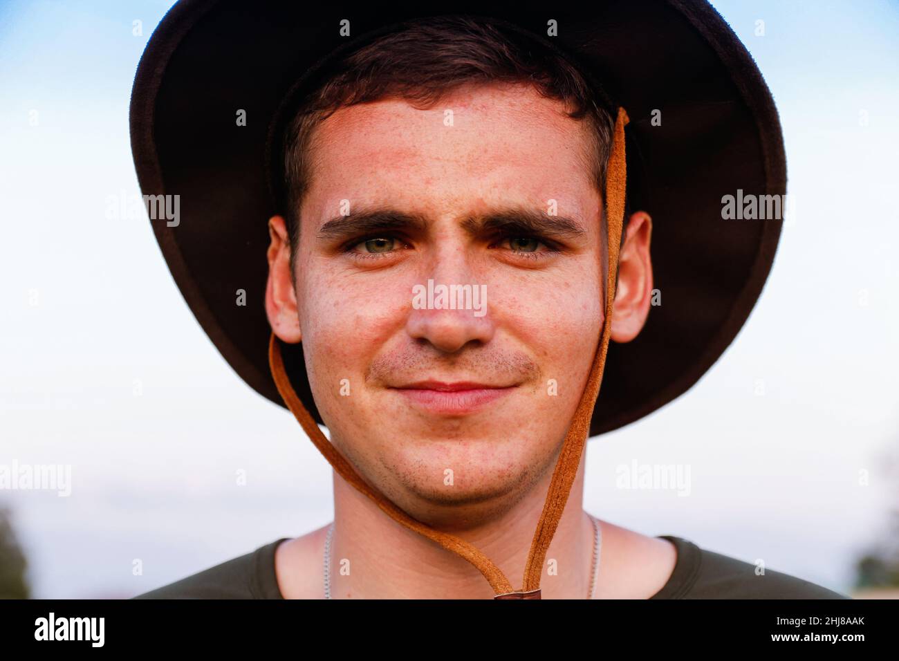 Junger Mann lächelnder Bauer im Cowboyhut auf dem landwirtschaftlichen Feld vor blauem Himmel. Porträt eines tausendjährigen Mannes, der im Freien auf dem Hintergrund der Natur steht. Lief Stockfoto