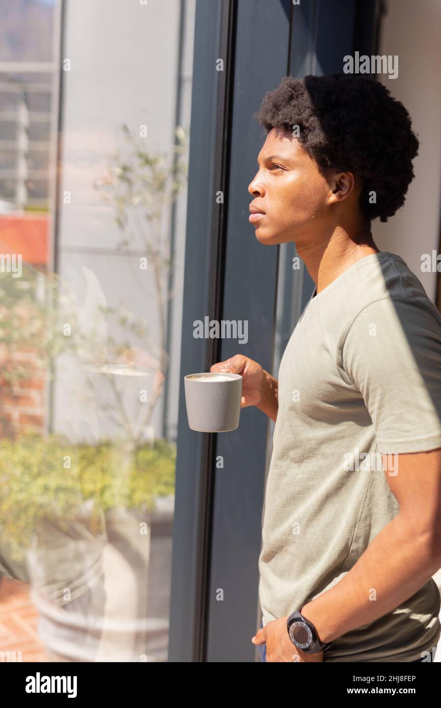 Seitenansicht eines nachdenklichen afroamerikanischen jungen Mannes mit Kaffeetasse, die zu Hause am Fenster steht. Unverändert, Lebensstil, Getränk, Kontemplation, Afro-Frisur Stockfoto