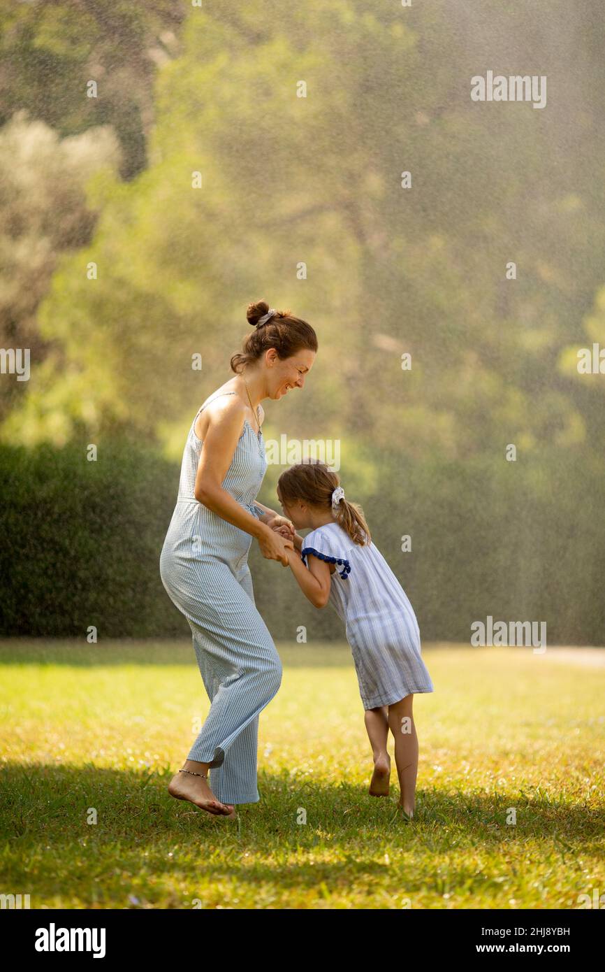 Nettes kleines Mädchen, das mit ihrer Mutter Spaß mit Wasser unter Bewässerungsregner hat Stockfoto