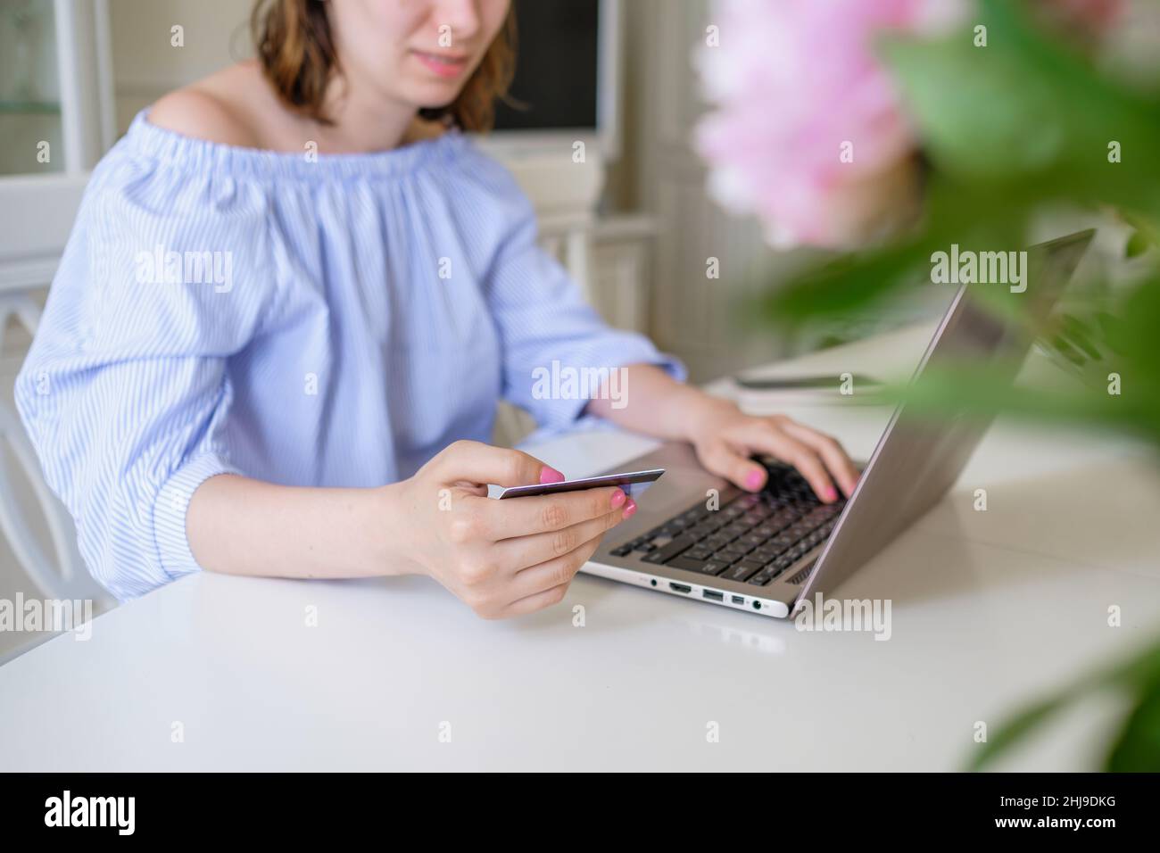 Die junge Frau in stilvoller Bluse kauft online über den Laptop und gibt Bankkartendetails hinter Blumen im Vordergrund zu Hause aus der Nähe ein Stockfoto