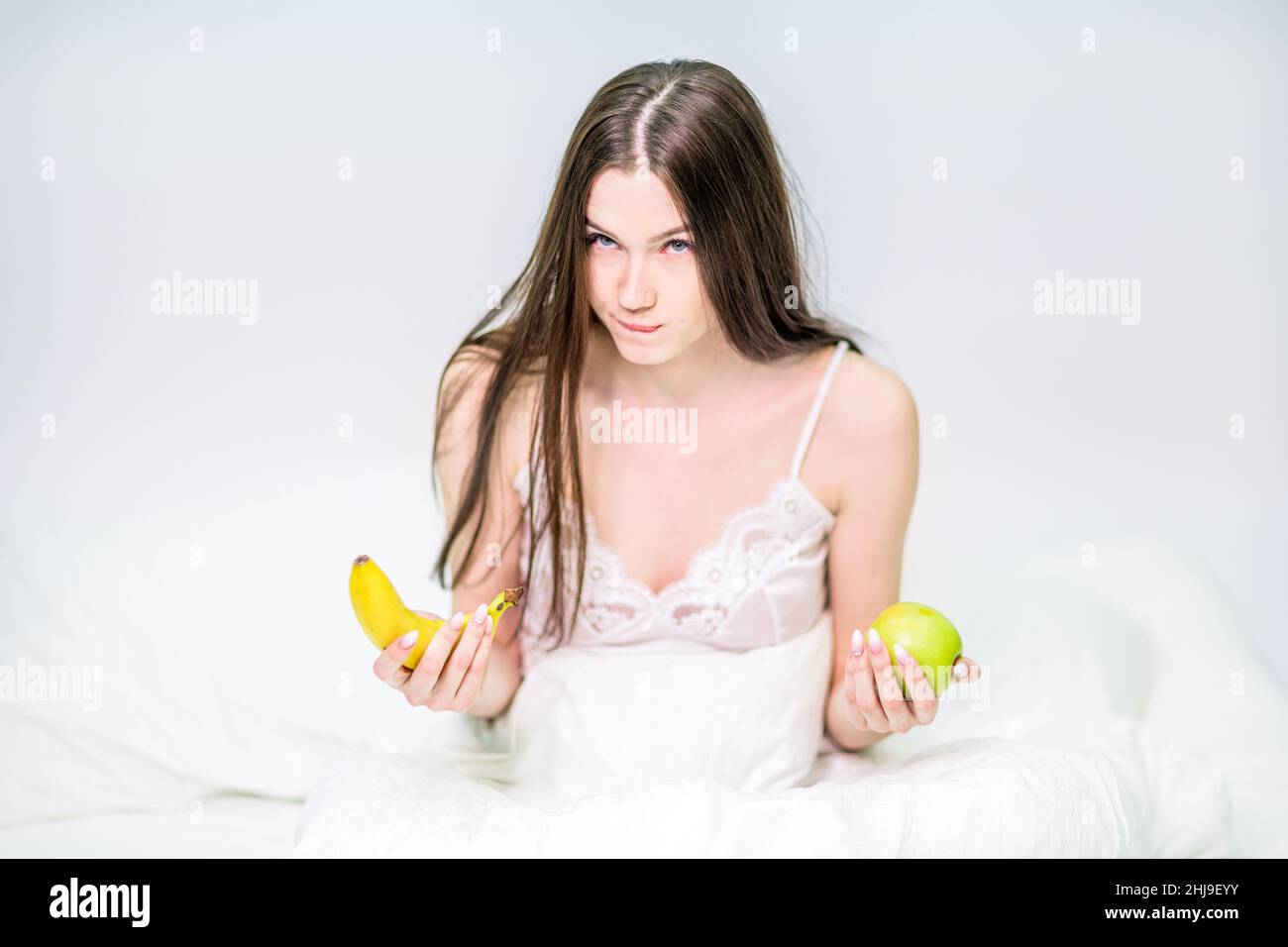Das Mädchen sitzt auf dem Bett, in eine Decke gehüllt, hält eine Banane und einen Apfel, wählt, was zu essen ist. Die Brünette frühstückte mit Obst. Stockfoto