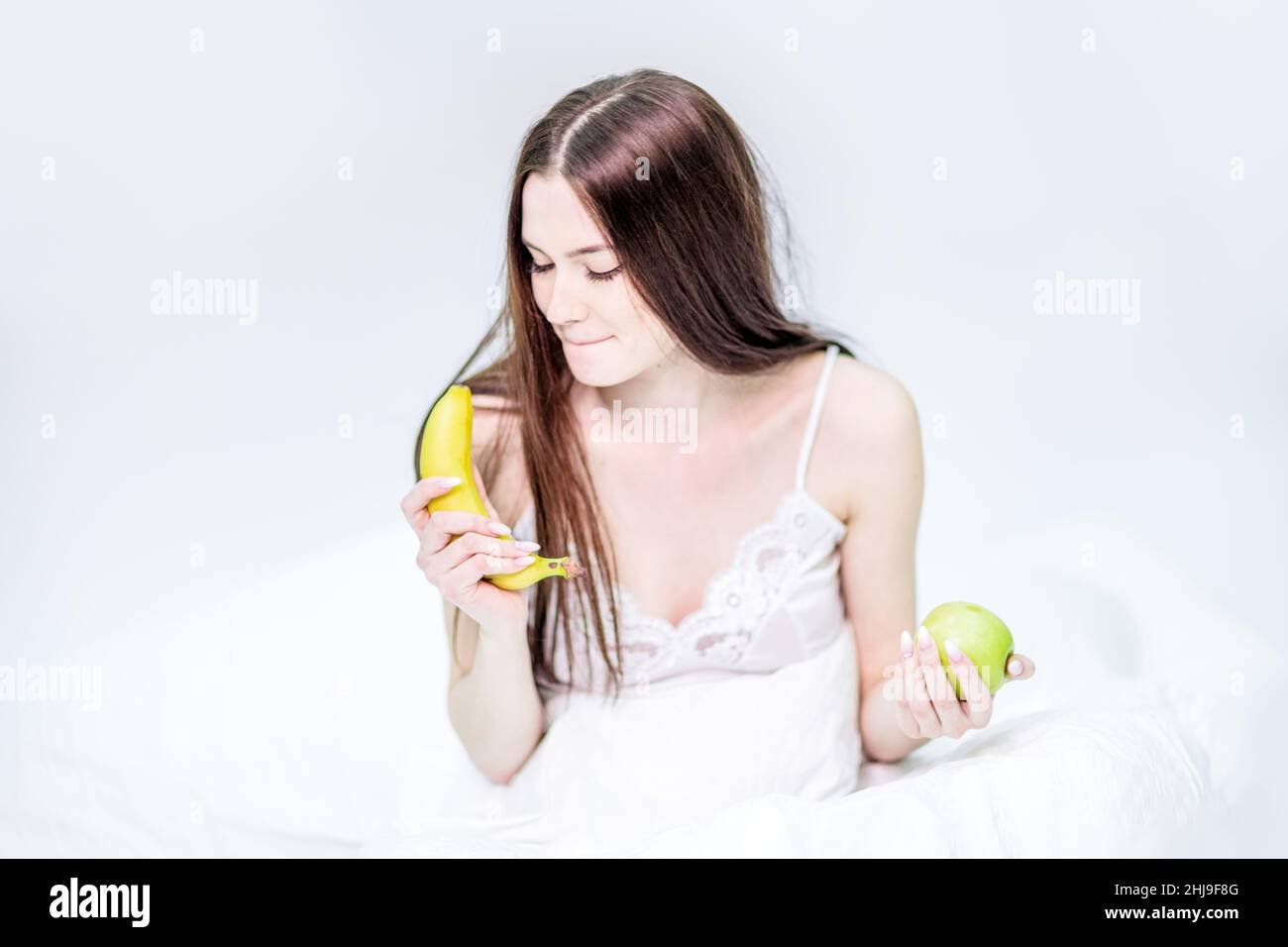 Das Mädchen sitzt auf dem Bett, in eine Decke gehüllt, hält einen Apfel und schaut auf eine Banane, wählt, was zu essen ist. Die Brünette frühstückte mit Obst Stockfoto