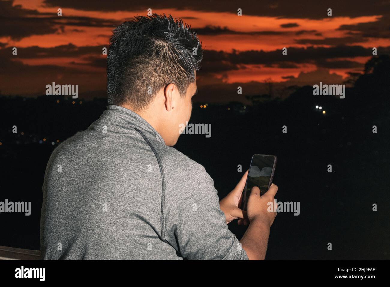 Junger braunhäuter lateinmann, der sich seine sozialen Netzwerke ansieht, im Hintergrund ein wunderschöner Sonnenuntergang in Colombia-Pereira, für den ein Student ein Foto machte Stockfoto