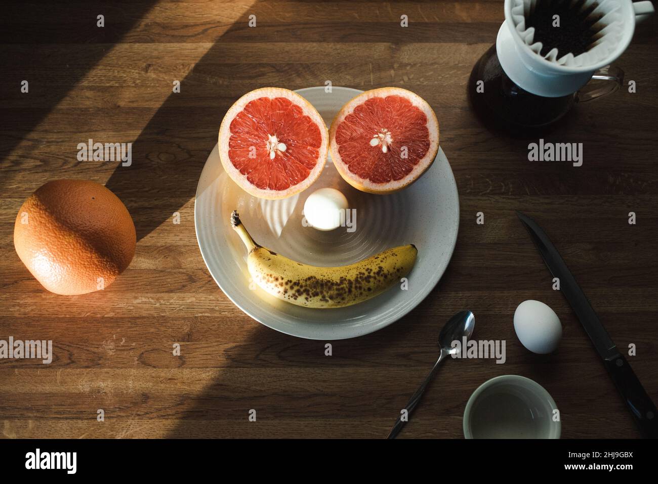 Eine Grapefruit, ein Ei und eine Banane bilden ein Frühstückslächeln auf einem weißen gesprenkelten Teller, umgeben von Utensilien, Kaffee in einem chemex und anderen Früchten Stockfoto