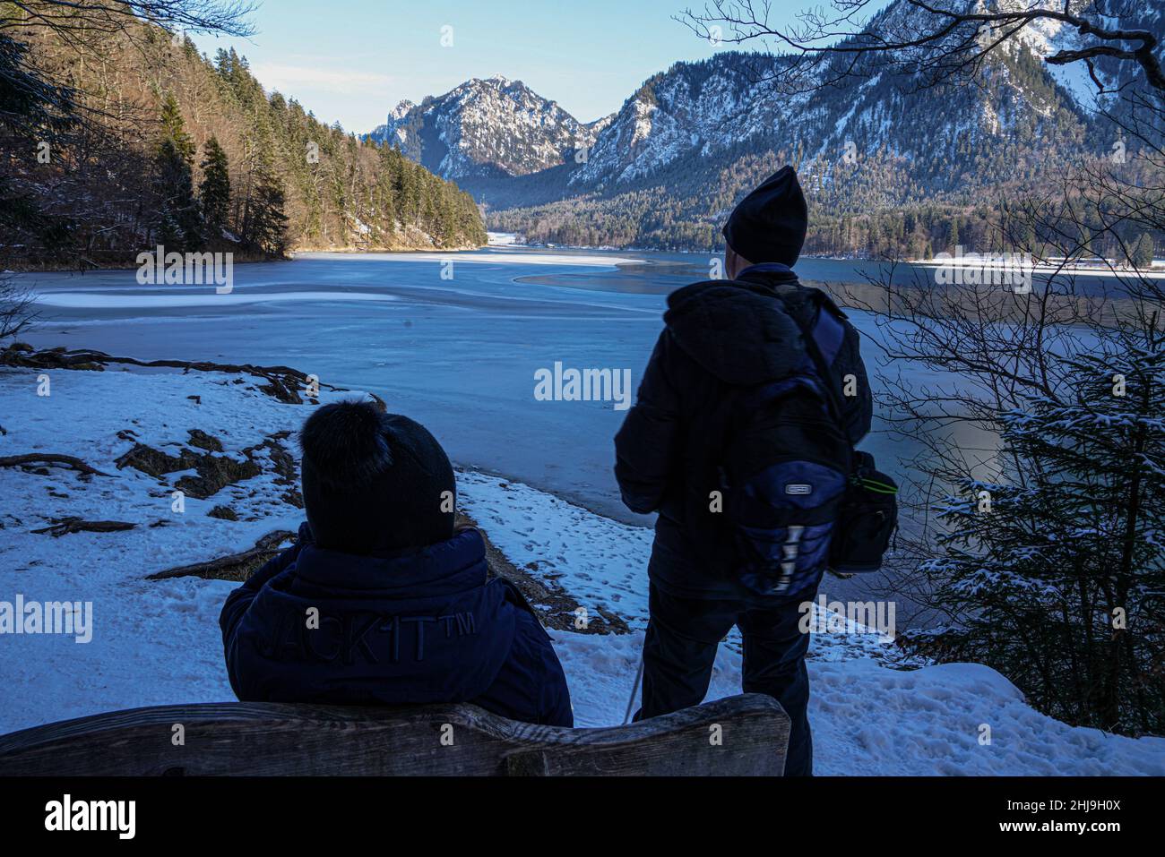 Ein Paar im Winter an einem Bergsee in den Alpen. Die Frau sitzt auf einer Bank und ruht sich aus. Der Mann steht und schaut auf die Berge. Stockfoto