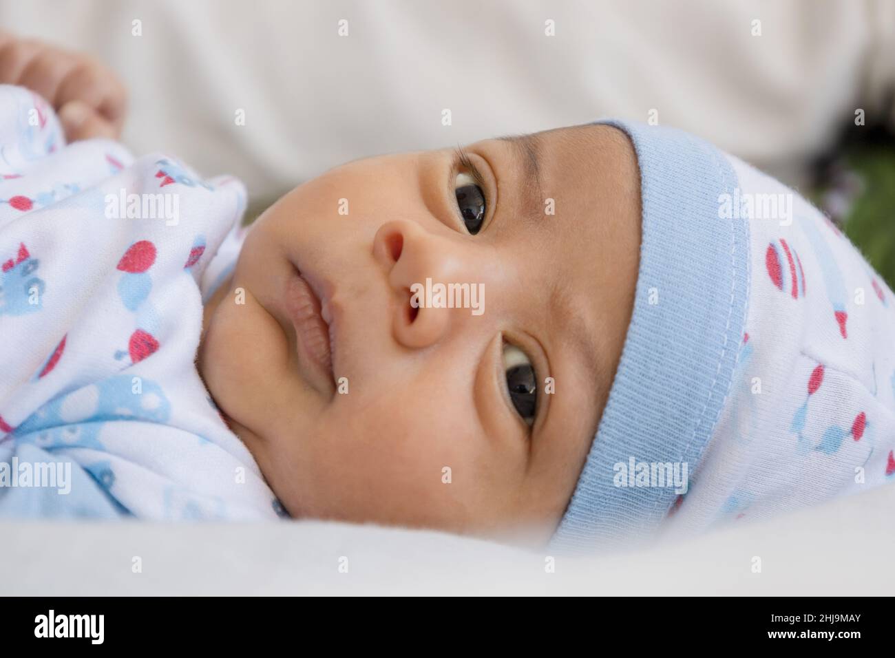 Das Gesicht eines zarten und schönen lateinischen Babys mit offenen Augen und glücklicher Ausstrahlung, das mit einem Hut auf seinem Bett liegt, einen neugeborenen Lebensstil, ein neues Leben und ein Kleinkind Stockfoto