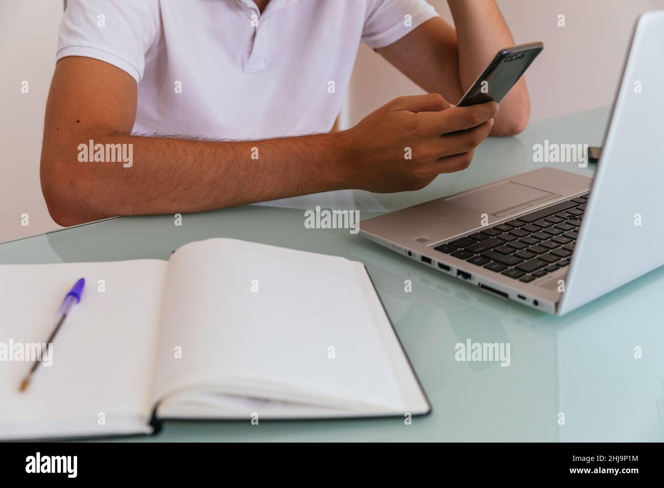 Seitenfoto eines nicht erkennbaren jungen Mannes im weißen Poloshirt, der zu Hause mit Smartphone in der Hand, Laptop und Notizblock arbeitet Stockfoto