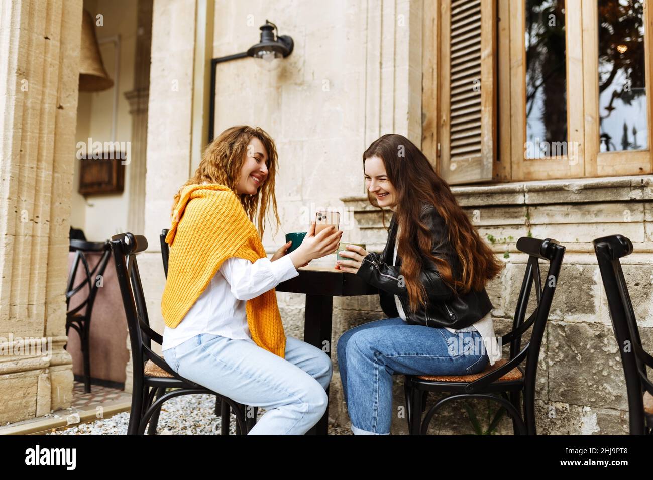 Fröhliche weibliche beste Freundin teilen Multimedia-Dateien auf Smartphones verbringen Freizeit zusammen, lächelnde Hipster Mädchen trinken Kaffee, sitzen auf der Terrasse Café. Stockfoto