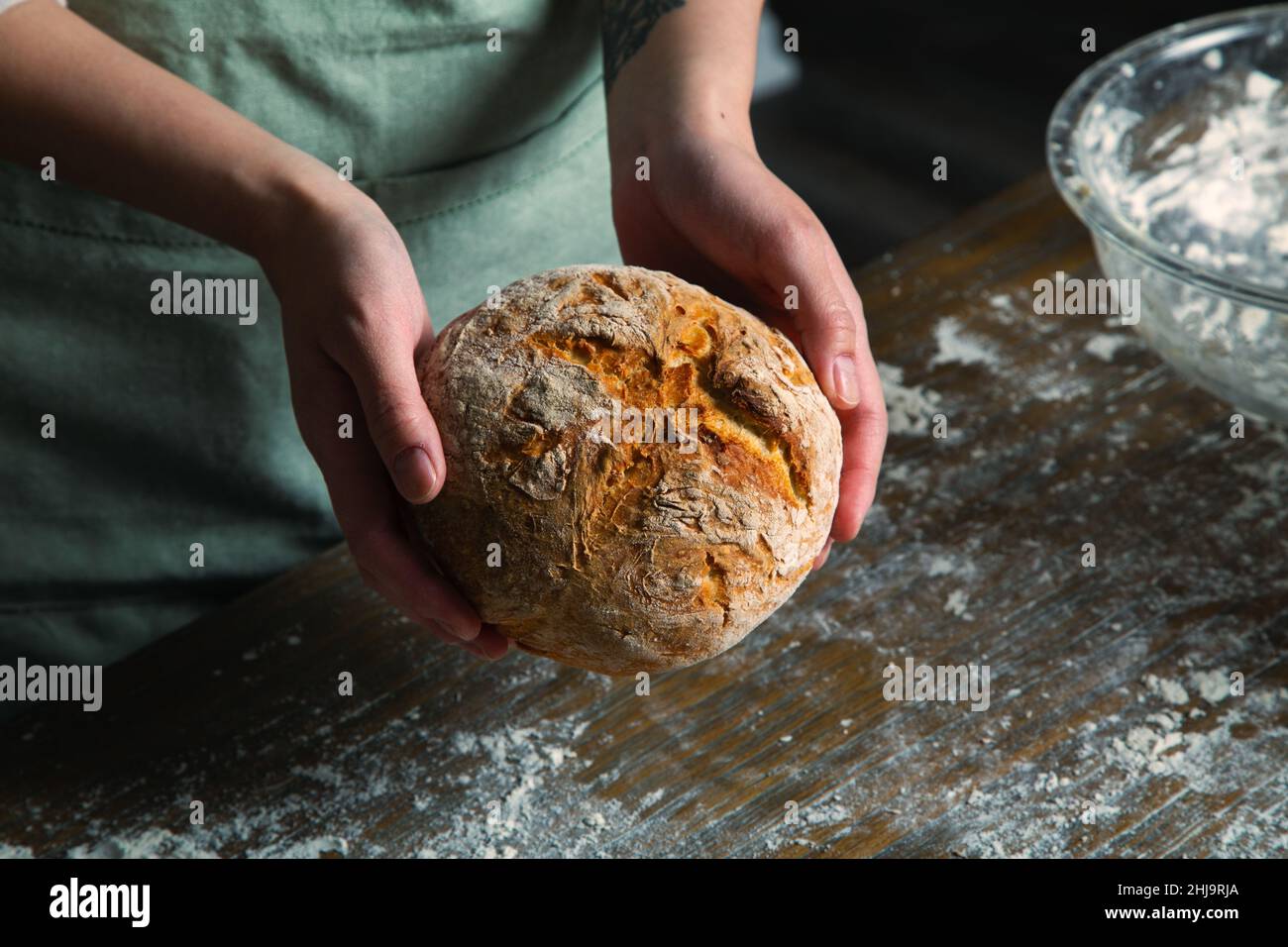 Ein Bild von einer Bäckerin in einer Küche, die einen frisch gebackenen, handwerklichen Brotlaib horizontal in den Händen hält. Stockfoto